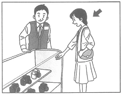 Scene: In a shop, a female customer is pointing at items inside a display case, while a male clerk behind the counter watches. An arrow points to the female customer — from the JLPT N5 July 2013 listening section